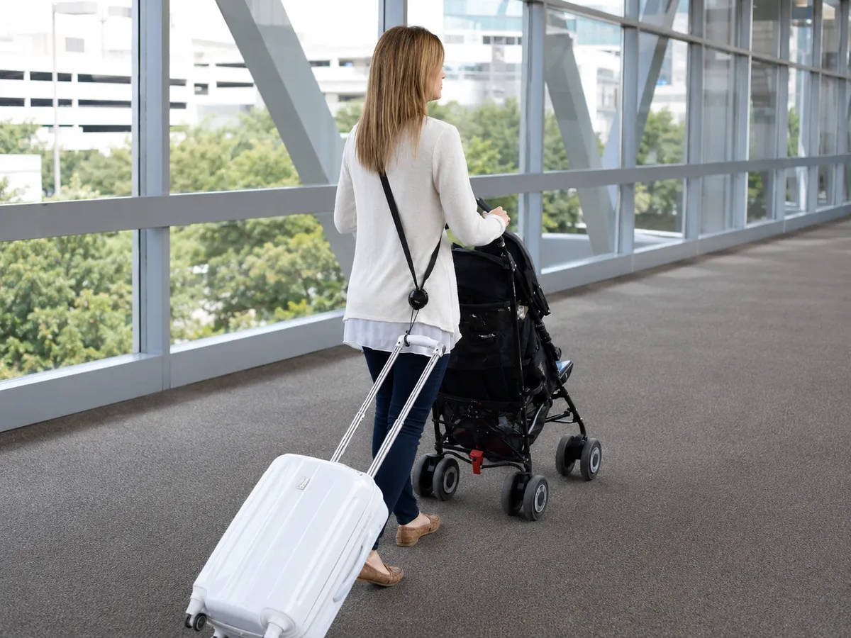 Parent pushing a stroller through an airport while a rolling carry-on follows behind using a RetraStrap luggage strap.