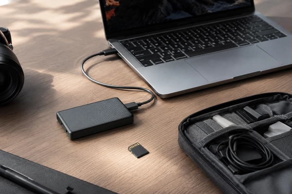 Portable SSD connected to a laptop beside a camera, memory card, and organized tech pouch
